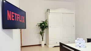 A hotel room with a bed, a TV displaying the Netflix logo, a white wardrobe, a potted plant, and neatly folded towels on the bed.