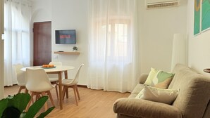 A living room with a beige sofa, a TV mounted on the wall, a dining table with chairs, and a window with sheer curtains.