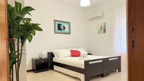 A bedroom with a bed, bedside tables, a nightstand, a potted plant, and a wall-mounted air conditioner.