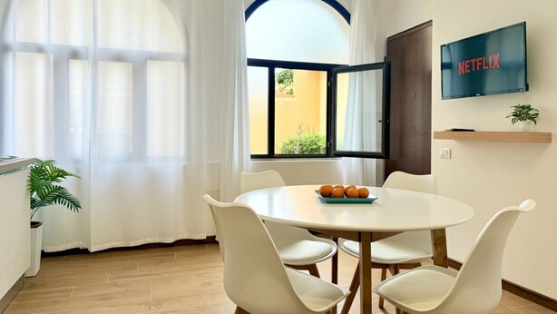 A dining area with a round table, white chairs, a fruit bowl, a TV on the wall, and a window with curtains.