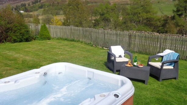 A hot tub in a backyard with two wicker chairs and a table, surrounded by a wooden fence and greenery. - Groose Hoose -  a cottage that sleeps 4 guests  in 2 bedrooms (Glenshee)