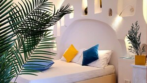 Ein Bett mit weißer Bettwäsche und zwei Kissen, eines gelb und eines blau, in einem Zimmer mit weißen Wänden und einer gewölbten Decke.