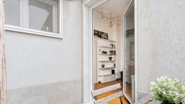A narrow outdoor corridor with a wooden bench, a potted plant, and a glass door leading to an interior space with shelves and a hanging light fixture. - Hostmate - Milano - Piazza Mentana (Milan)