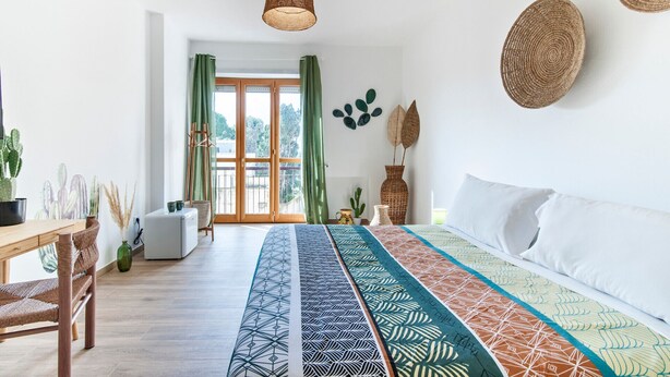 A modern bedroom with a large bed, a wooden desk, and a chair. There are two hanging wicker lamps, a green curtain, and a view of the outside through a glass door. - 7Rooms - The Countryside (Lecce)