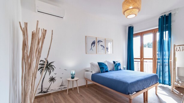 A bedroom with a wooden bed, a small bedside table, a wall-mounted air conditioner, a hanging light fixture, and a window with curtains.