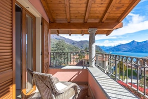 Un balcon avec un fauteuil en osier, une colonne en pierre et une vue sur les montagnes et un lac.