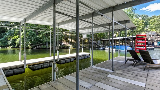 Une jetée abritée, dotée de chaises et d’un banc rouge, surplombe une étendue d’eau, le tout étant entouré d’arbres et avec une maison visible au loin.