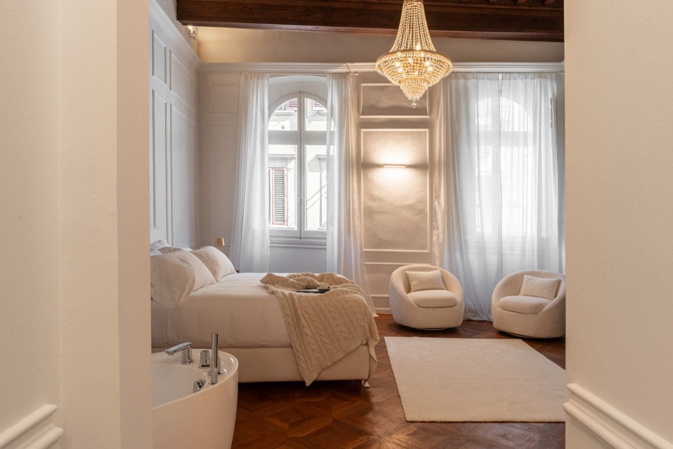 A bedroom with a bed, a chair, a chandelier, and a window with curtains.