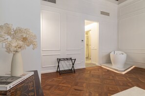 A room with a wooden parquet floor, a white door, a white vase with dried flowers, and a black metal rack.