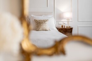 Una cama bien hecha con sábanas blancas y un cojín estampado, una mesita de noche con lámpara, y una pared con molduras decorativas.