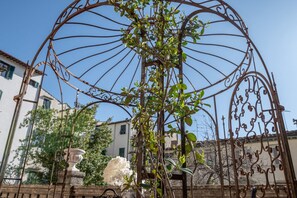 Una estructura de hierro forjado con una planta trepadora, un jarrón de piedra y un edificio al fondo.