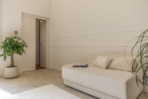 Ein neutral gestaltetes Wohnzimmer mit einer Couch, einem Blumentopf und einer Tür zu einem anderen Raum.