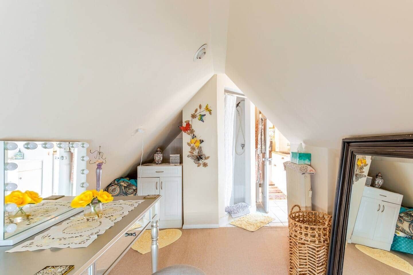 A cozy attic bedroom with a vanity, a mirror with lights, a bed with a decorative headboard, and a view of the living area.