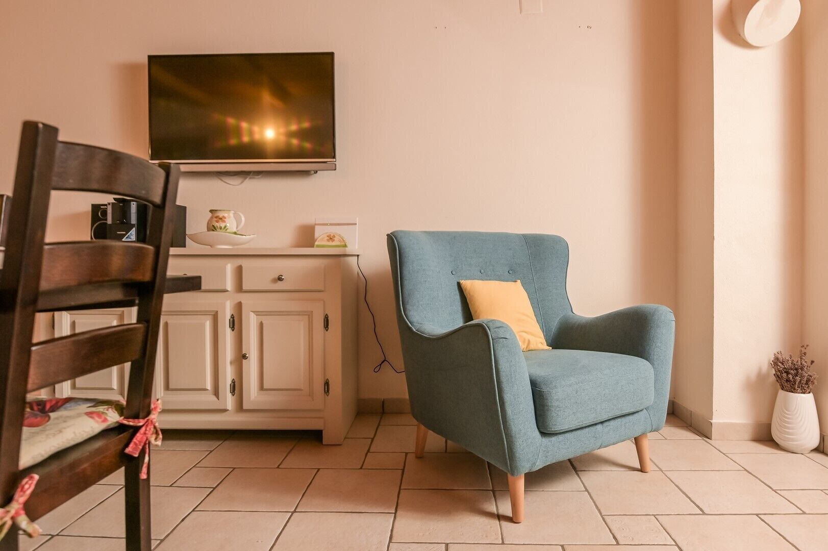 Un salon avec un fauteuil bleu, une chaise en bois, un meuble et un téléviseur à écran plat fixé au mur.