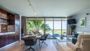 Un salon moderne comprenant un canapé, une table à manger avec des chaises, un téléviseur fixé au mur et une grande porte-fenêtre coulissante donnant sur un espace extérieur agrémenté de plantes.