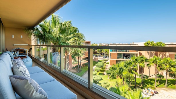 Un balcon aménagé avec un canapé, une table à manger et des chaises, donnant sur un espace paysager agrémenté de palmiers et d’une piscine.
