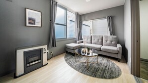 A modern living room with a grey sofa, a round coffee table, and a large window with curtains. - VITA (Furano)