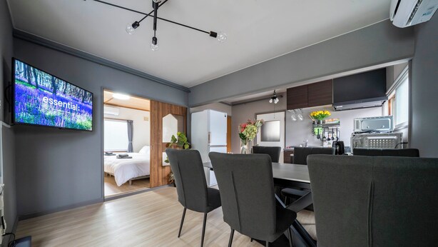A modern kitchen with a flat-screen TV, a dining area with chairs, and a visible bedroom with a bed. - VITA (Furano)