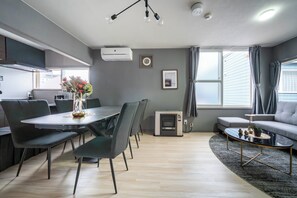 A modern living room with a dining area, grey walls, a grey sofa, a coffee table, and a large window with curtains. - VITA (Furano)