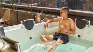 A man sitting in a hot tub with a drink, surrounded by a window view and a small table with a bottle and grapes. - Rosa Negra Stadio Aparta-suite Your Cozy Retreat (Medellín)