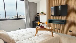 A hotel room with a large bed, a wooden TV stand, a small table with fruit and juice, and a city view. - Stadio Serenity Rosa Negra Aparta-suite (Medellín)