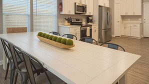 Uma cozinha moderna com armários brancos, eletrodomésticos de aço inoxidável e uma mesa de jantar comprida com cadeiras.