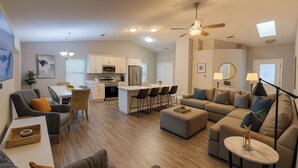 Uma sala de estar moderna com sofá, um banquinho e poltronas, uma área de jantar com mesa e cadeiras, e uma cozinha com armários e eletrodomésticos brancos.