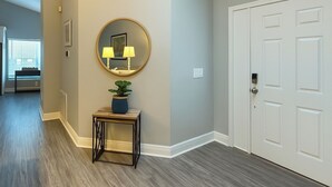 Um corredor com piso de madeira, um espelho redondo com duas luminárias, uma pequena mesa com uma planta e uma porta branca.