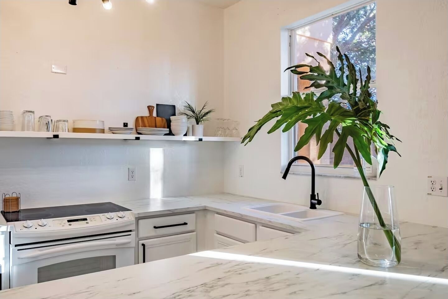A modern kitchen with white cabinetry, a built-in stove, a sink, and a large window providing natural light.