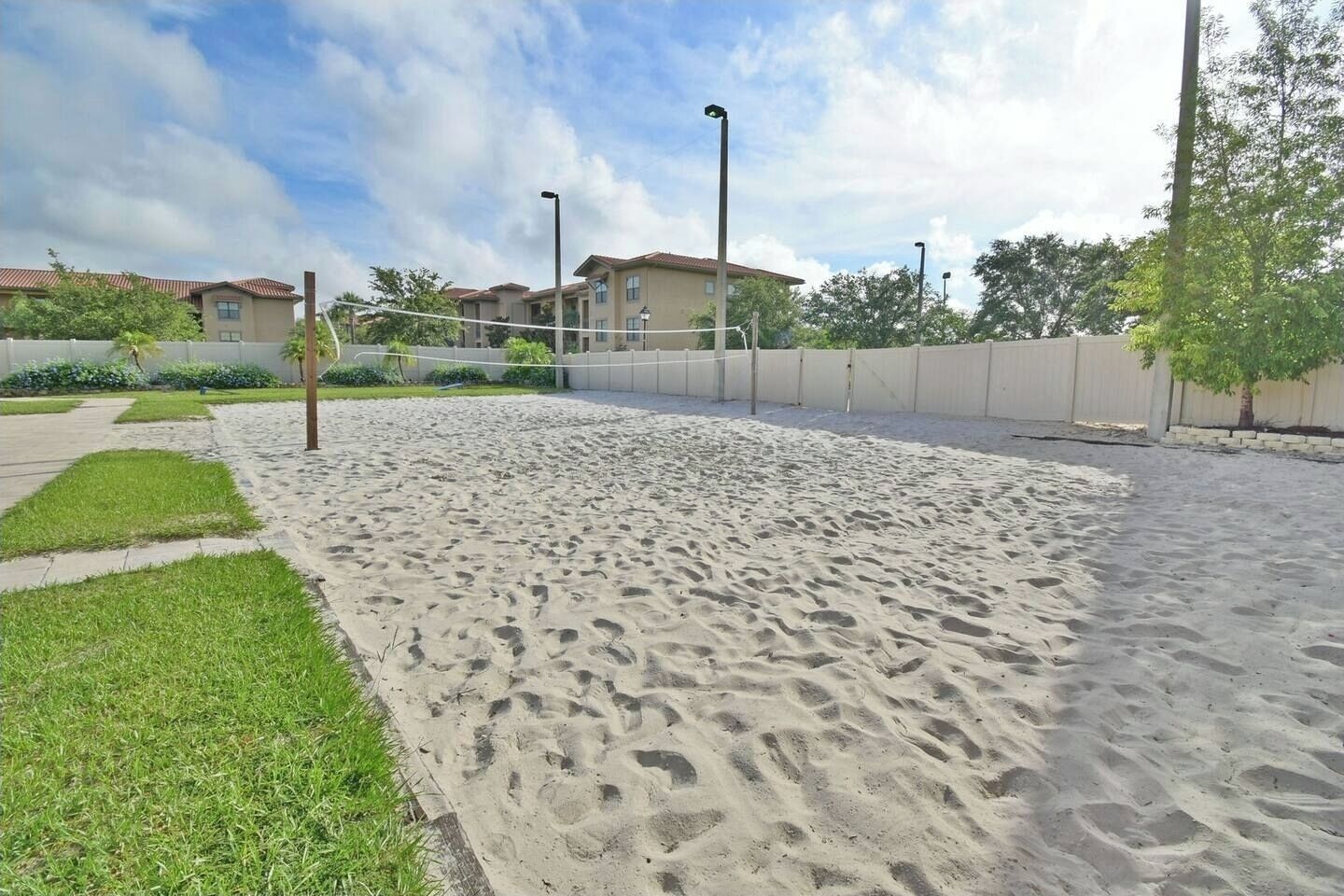 A sandy outdoor area with a volleyball net, surrounded by a low fence and residential buildings in the background.