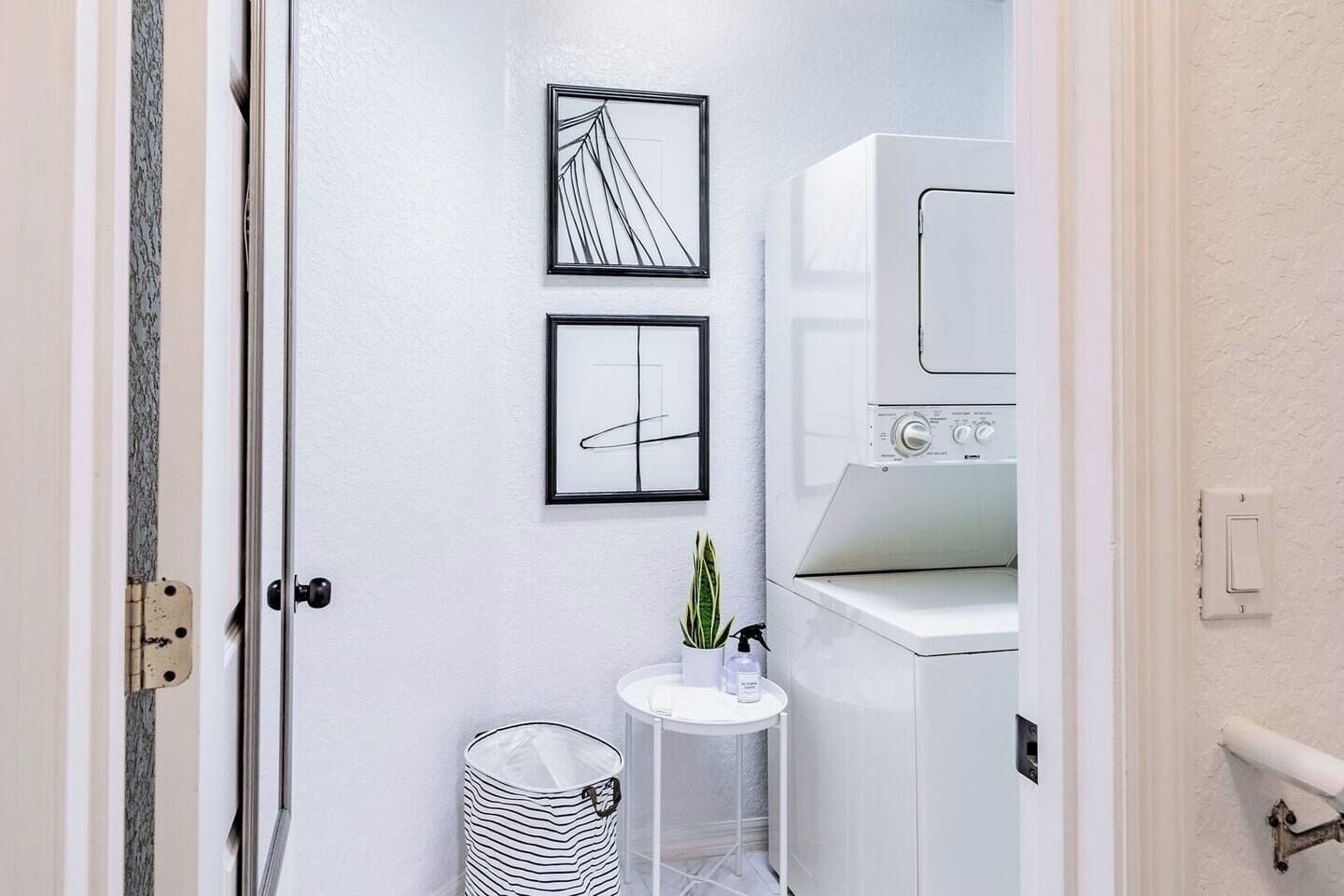 A laundry room with a washing machine, two framed abstract artworks, a small round table with a potted plant, and a striped trash bin.