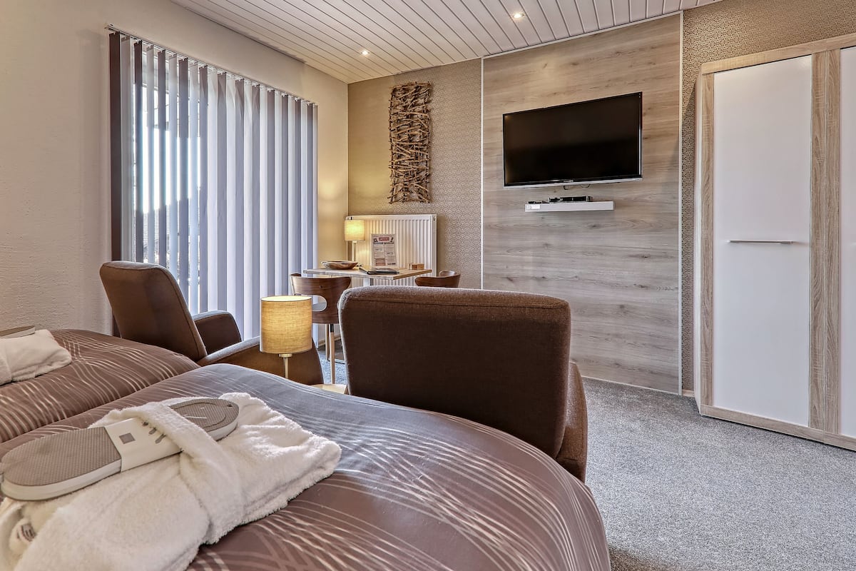 A hotel room with a large bed, two chairs, a desk with a lamp, a flat-screen TV mounted on the wall, and a wardrobe.