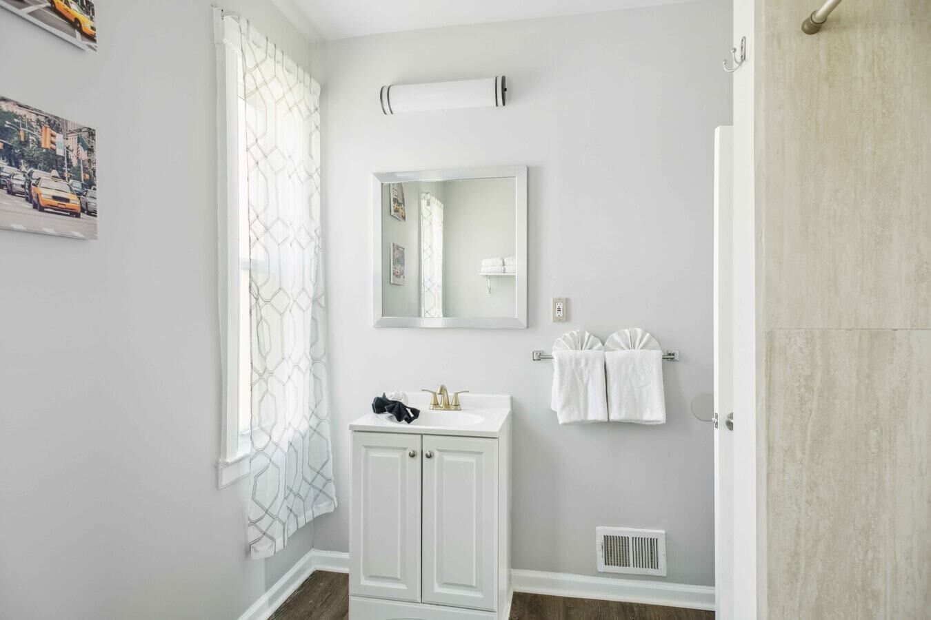 Une salle de bain avec un meuble-lavabo blanc, un miroir, un porte-serviettes avec deux serviettes et une fenêtre agrémentée d’un rideau à motifs géométriques.
