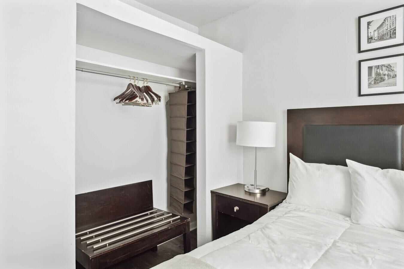 A hotel room with a bed, a bedside table with a lamp, a nightstand, a wooden bench, a closet with hanging clothes, and a framed picture on the wall.