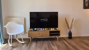 A modern living room with a flat-screen TV on a wooden TV stand, a white rocking chair, and decorative items. - Appt 80m², 13 P,  Disneyland, Fibre, 2 parkings (Chessy)