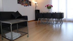 A modern living room with a black sofa, a small table with a wire basket, a standing lamp, and a dining area with chairs.