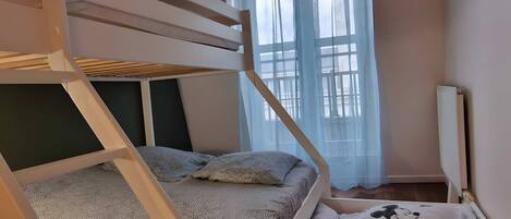 A bunk bed with a grey knitted blanket and pillows, a window with sheer curtains, and a Mickey Mouse pillow on the floor.