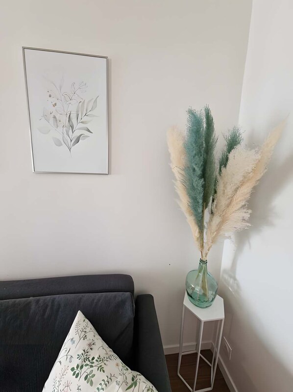 A living room with a dark sofa, a small white side table with a vase of pampas grass, and a framed botanical print on the wall.