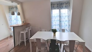 A dining area with a white table and chairs, a small plant on the table, and a window with floral curtains. - Gd 4P 85m² hyper centre Disney (Chessy)