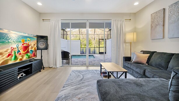 A living room with a large flat-screen TV, a grey sofa, a wooden coffee table, and a view of a swimming pool through glass doors. - Relaxing Nature Escapade with Game Room (Clermont)