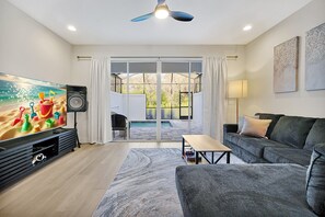 Uma sala de estar com uma televisão de tela plana grande, um sofá cinza, uma mesa de centro de madeira e vista para uma piscina através de portas de vidro.