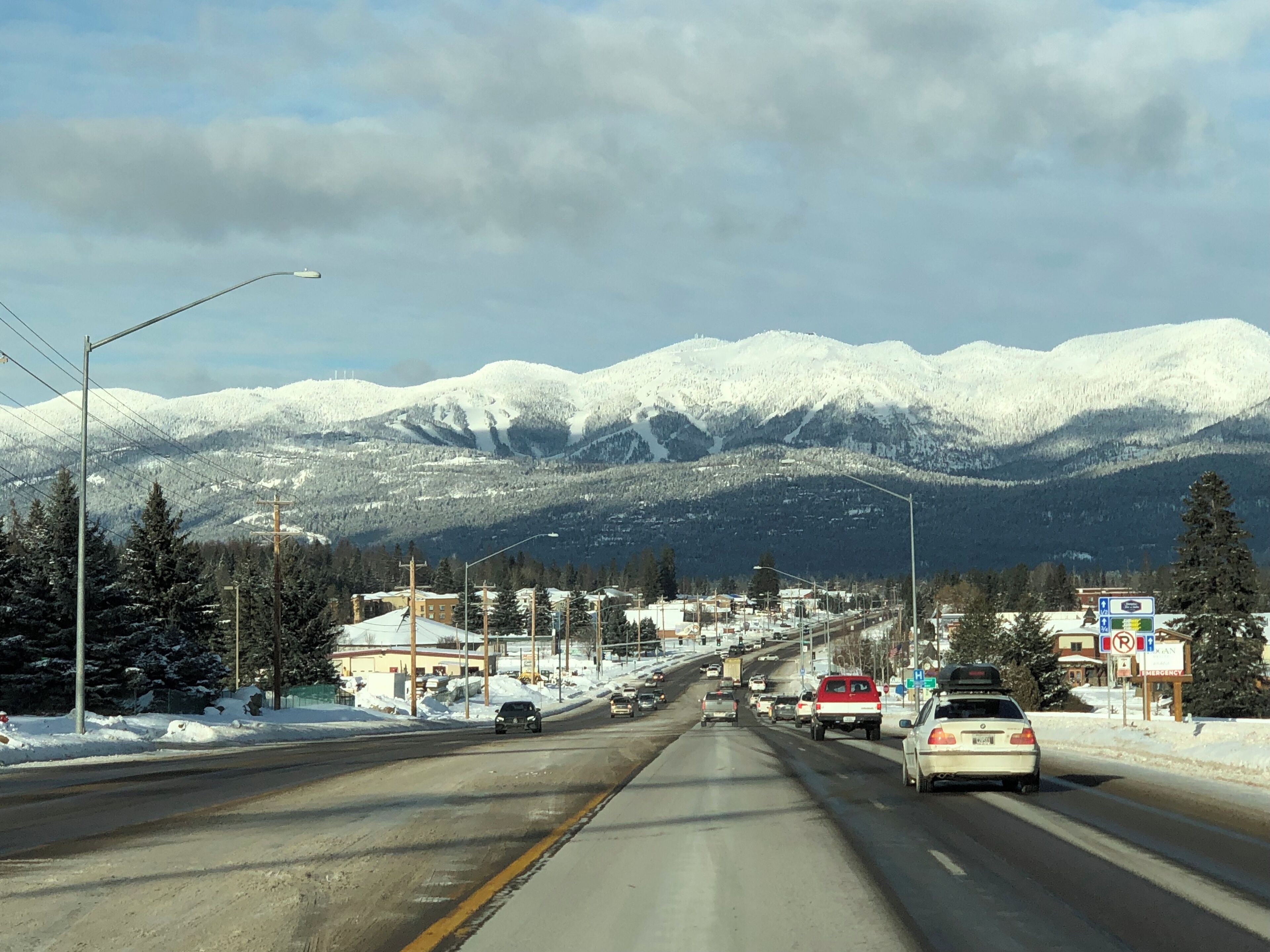 A road with vehicles, snow-covered mountains, and buildings in the background.