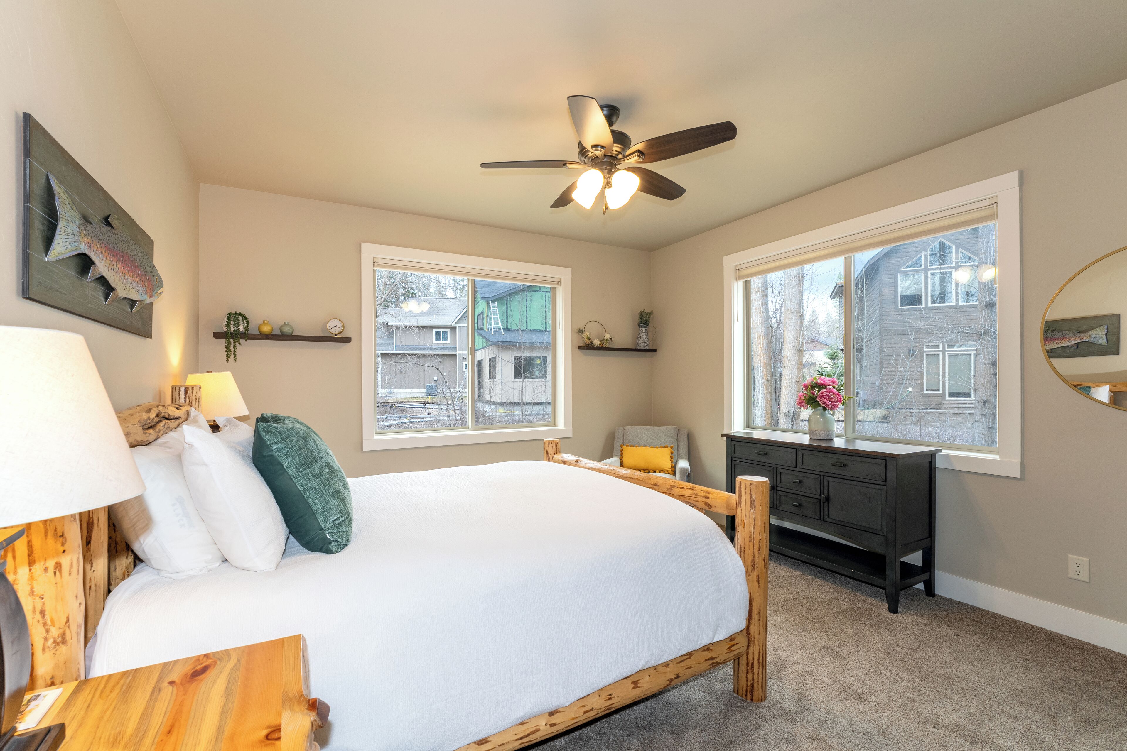 A bedroom with a bed, bedside table, lamp, ceiling fan, dresser, and large windows.