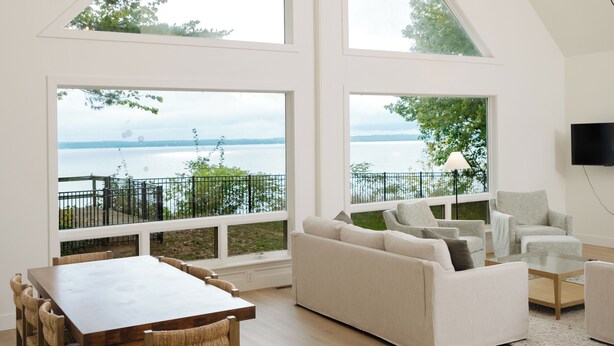 A spacious living area with a high ceiling, large windows offering a view of the outdoors, a dining table with chairs, and a seating area with sofas and a coffee table. - Luxury Waterfront Home in Suttons Bay (Suttons Bay)