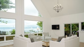 A spacious living room with large windows, a chandelier, a flat-screen TV, and a view of the water. - Luxury Waterfront Home in Suttons Bay (Suttons Bay)