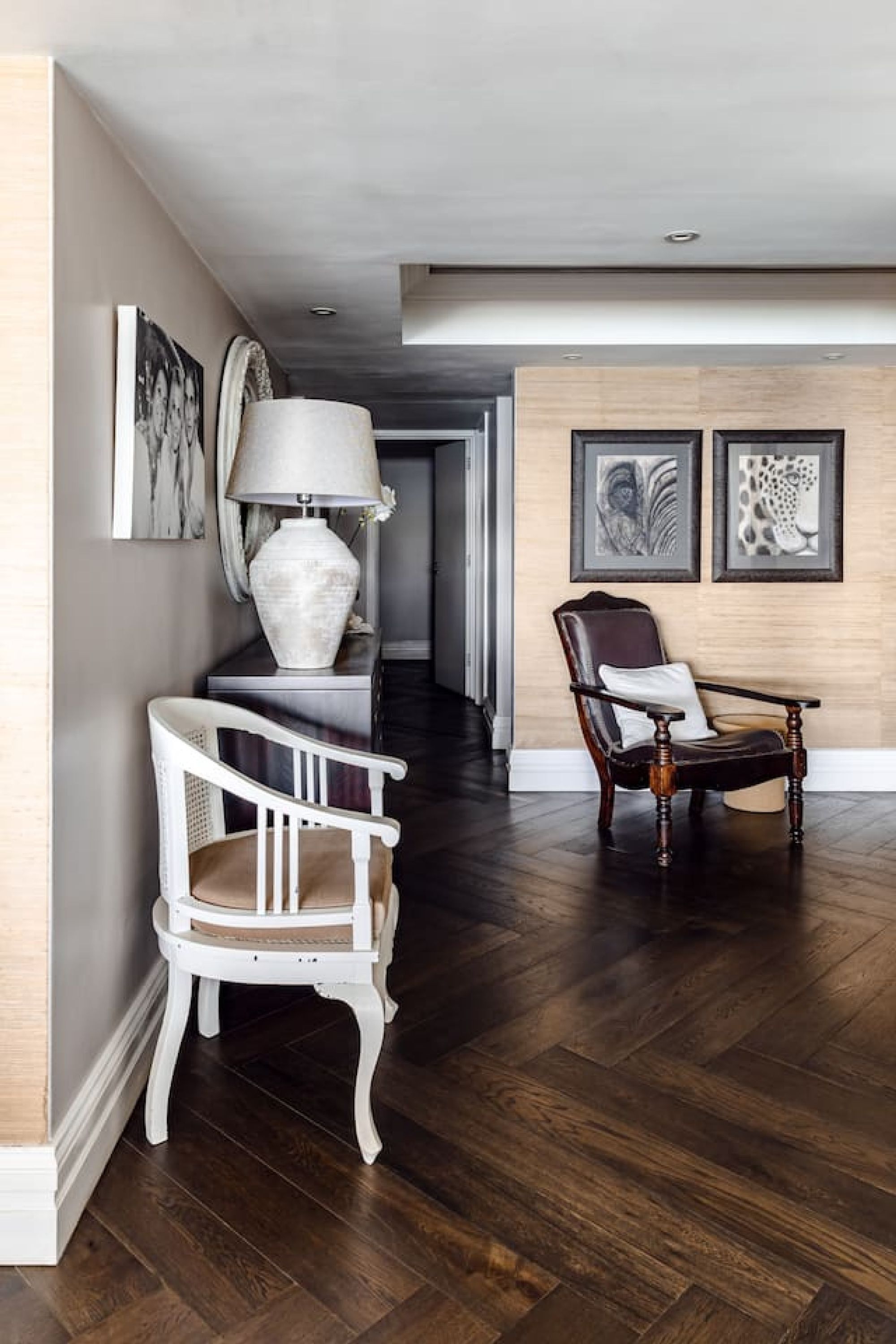 A hallway with dark wooden flooring, a white chair, a dark wooden chair, and a white vase on a console table.