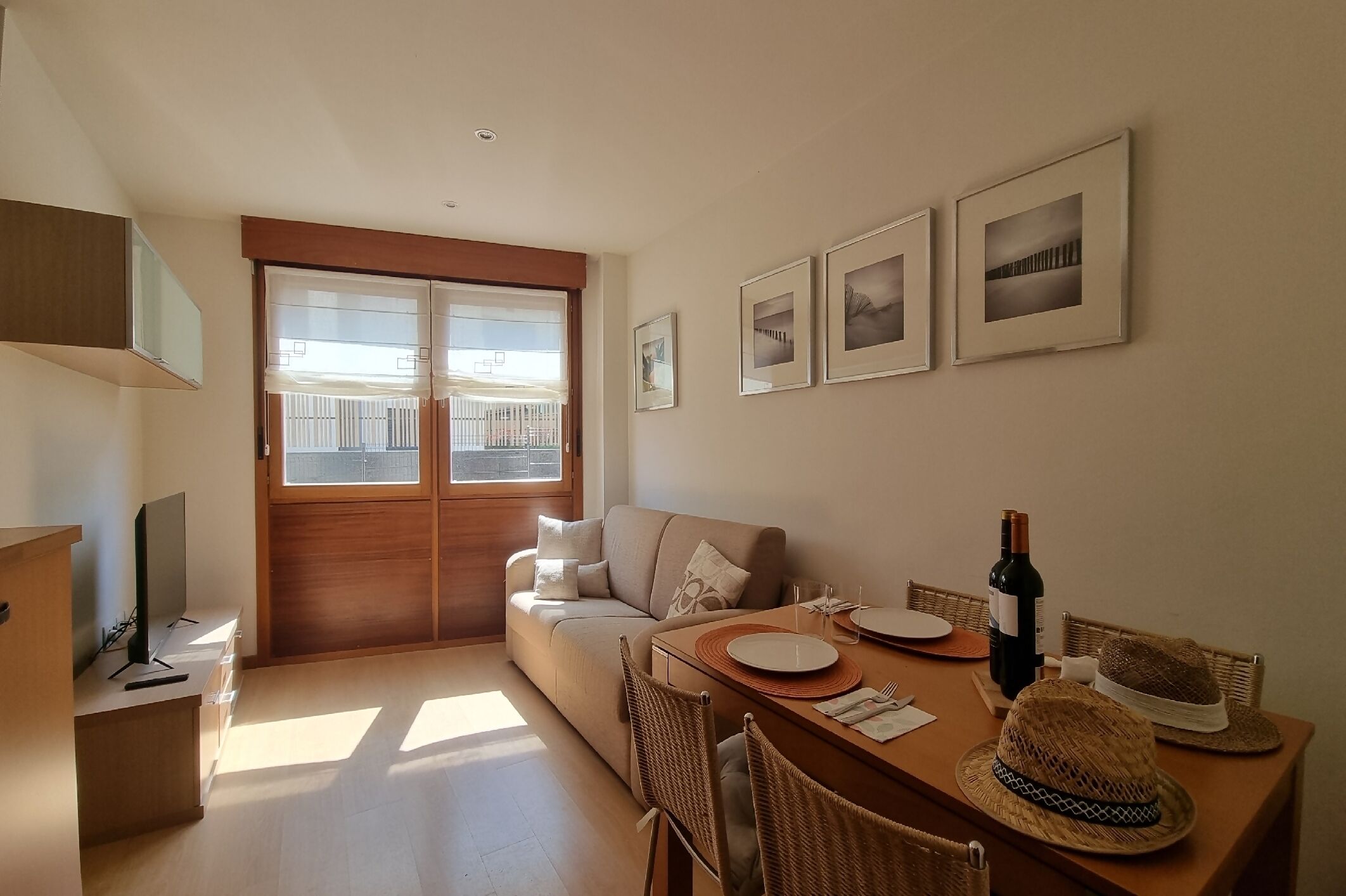 Een moderne woonkamer met een bank, eettafel en stoelen. Er hangen afbeeldingen in lijsten aan de muur, een raam met uitzicht en een tv op een dressoir.