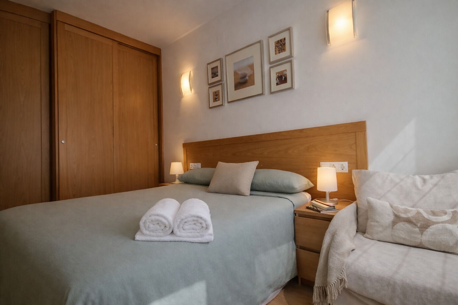 A neatly made bed with two rolled towels, a wooden headboard, bedside lamps, and a small bedside table with books and a lamp.