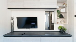 Un salon moderne avec un téléviseur à écran plat fixé à un mur blanc, un meuble de télévision en bois foncé et une petite étagère ornée d’objets décoratifs.