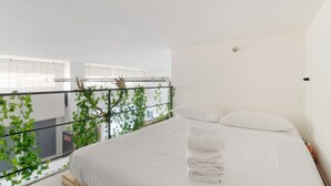 Une chambre à coucher avec un lit, du linge de lit blanc, une fenêtre avec des stores et un balcon agrémenté de plantes.